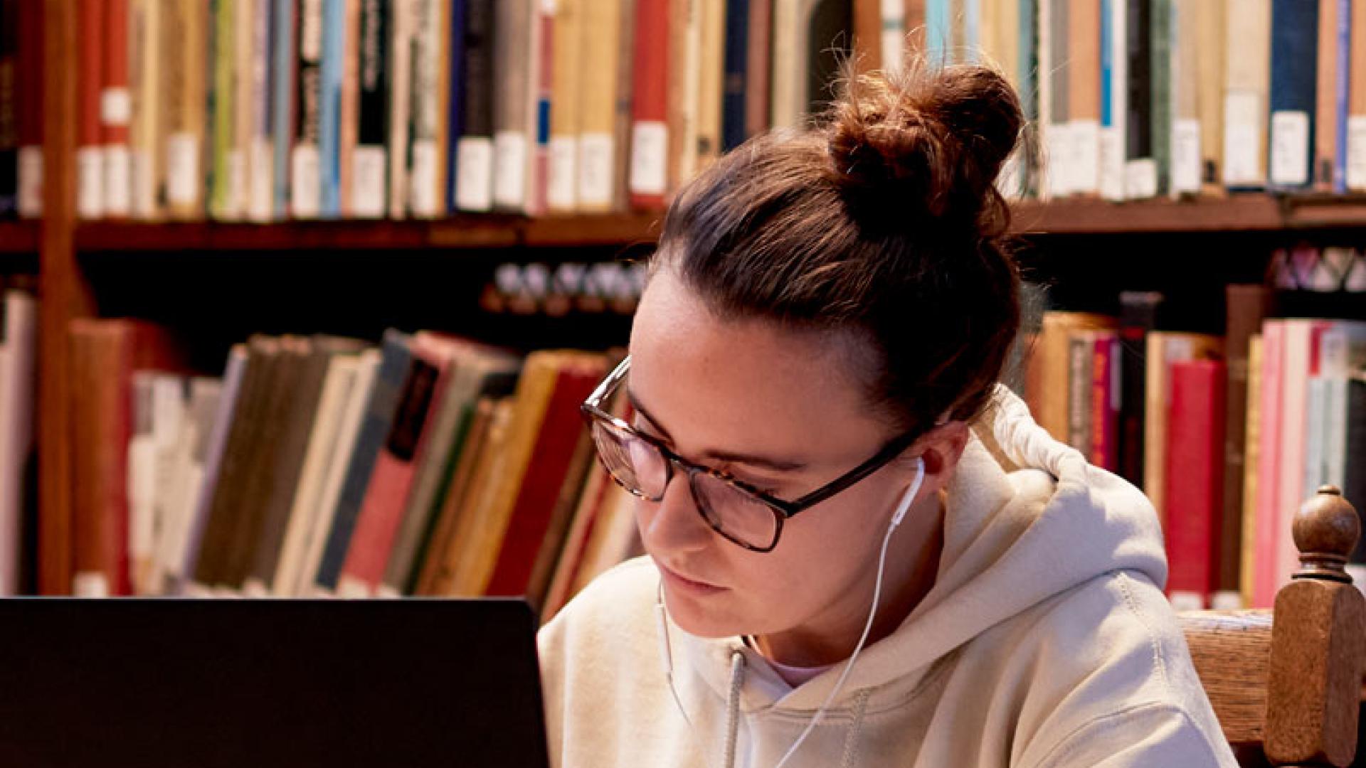 Student sitting in the library with a laptop and books on shelves in the background