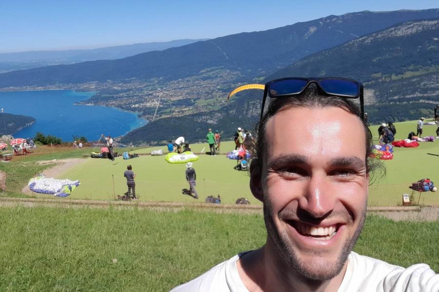 Marley Willegers smiles into camera with sunglasses onhis head, behind him are people preparing to paraglide and landscape with lake far below and beyond.